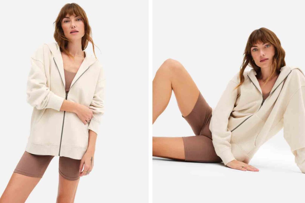 Model wearing a MATE the label white oversized hoodie with brown shorts. There are two images; on the left, she is standing. On the right, she sits on the ground, leaning to the right.