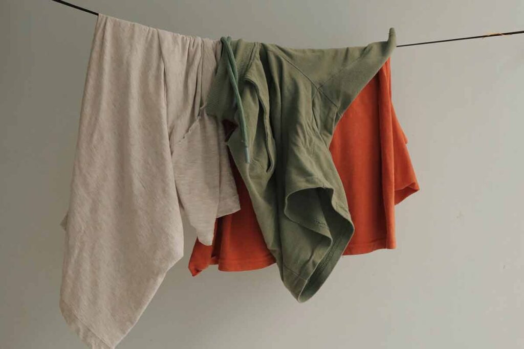 Organic natural fiber clothing hangs on a clothing line. There are three garments; in colors white, green, and orange.