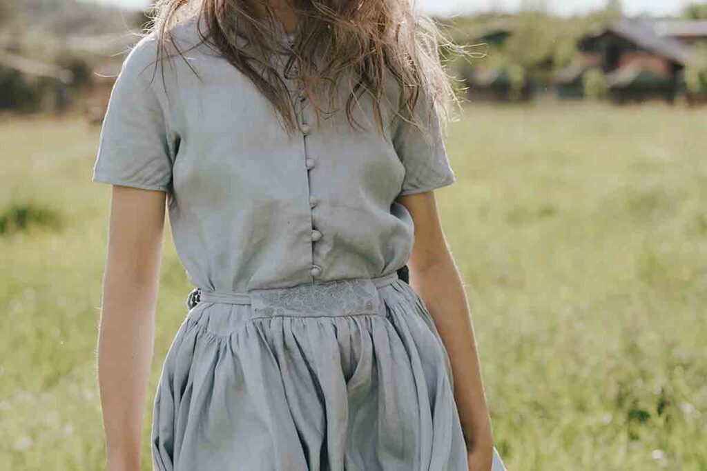 Woman stands in a grass field in a blue and gray organic natural fiber cotton dress. The image shows her from the neck down.
