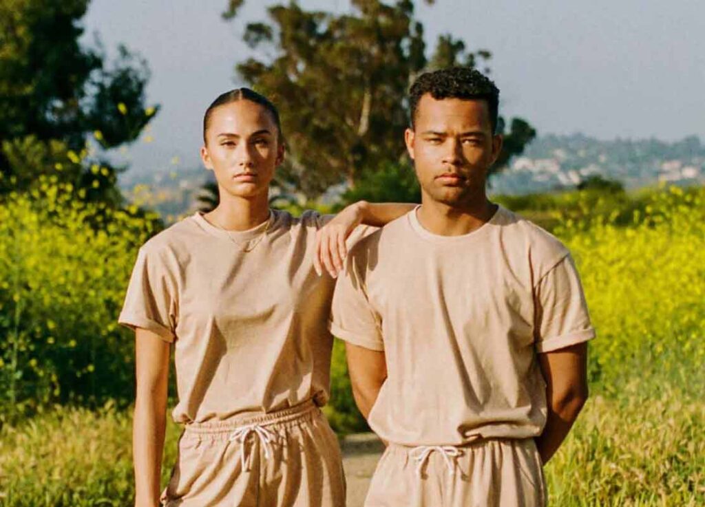 A man and a woman stand in a field wearing matching brown organic cotton sweat suits, including a t shirt and pants. They stand facing the camera, with the woman's arm on the man's shoulder, modeling one of the best organic clothing brand garments.