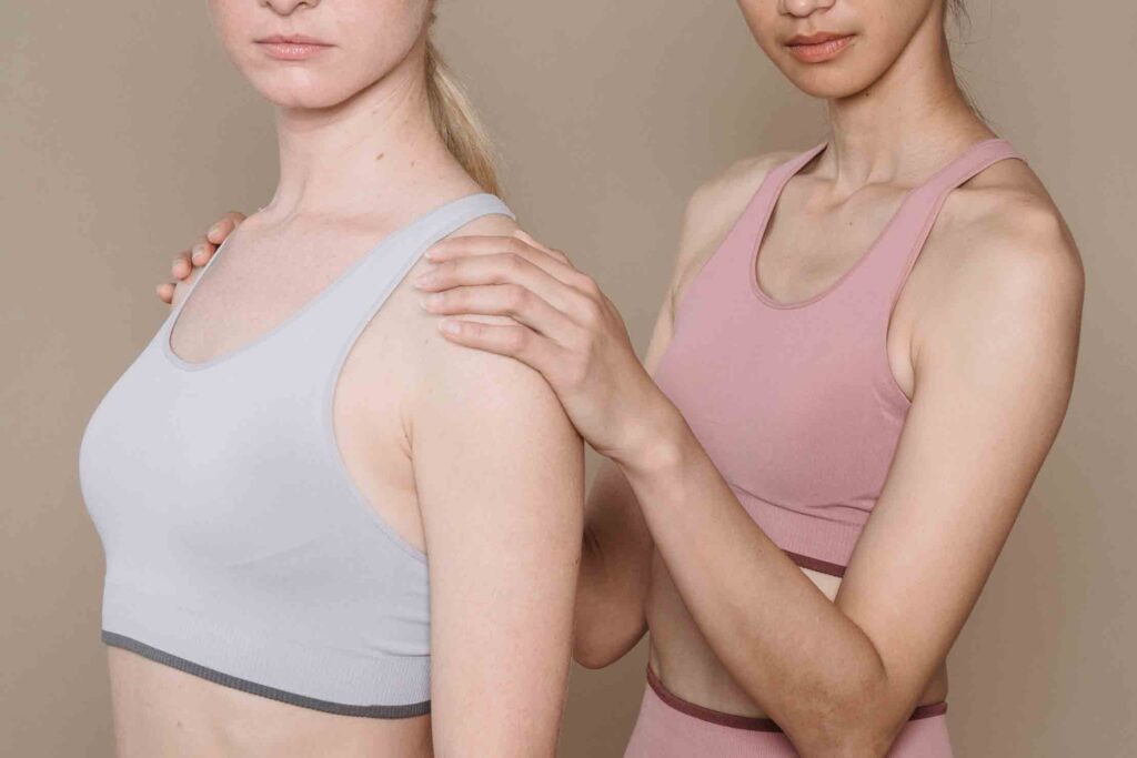 Two women wear synthetic clothing activewear. One wears a white nylon sports bra, the other a pink sports bra.