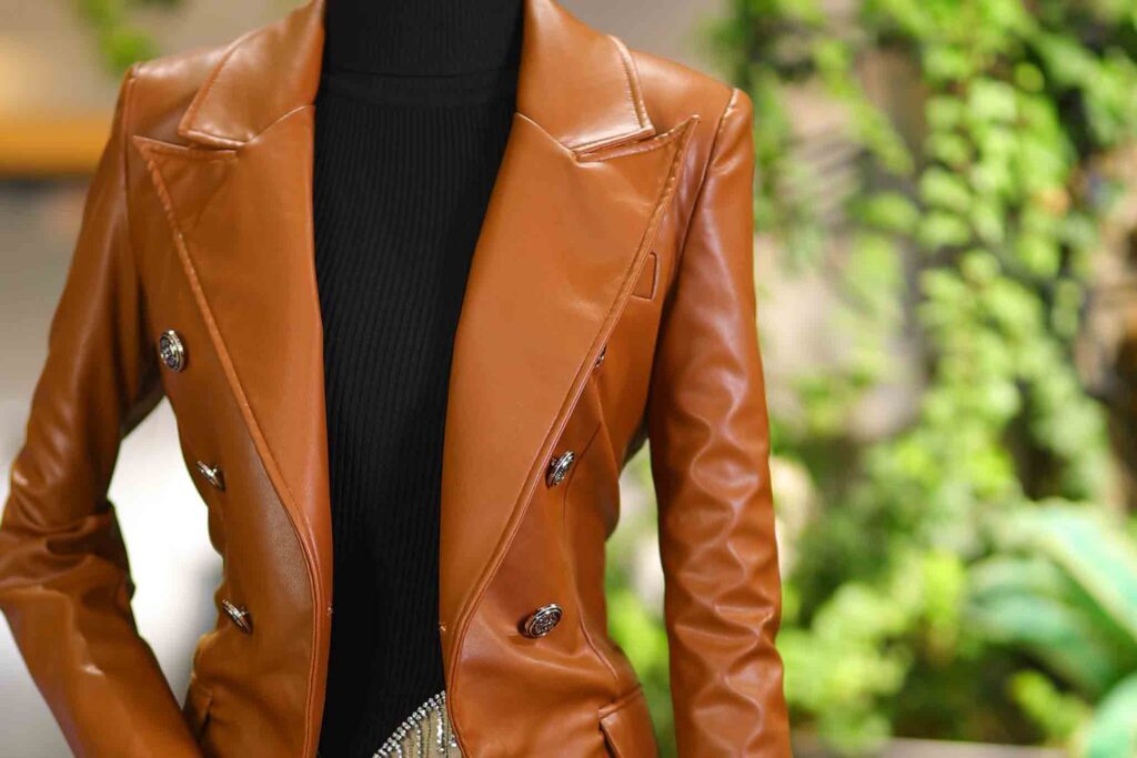 A mannequin wears a brown leather jacket and black undershirt. Green foliage is in the background. 