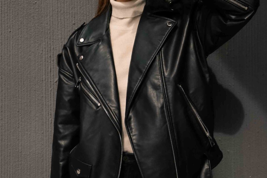 A woman wears a black leather jacket and a white undershirt. She stands against a black background with the jacket unzipped.