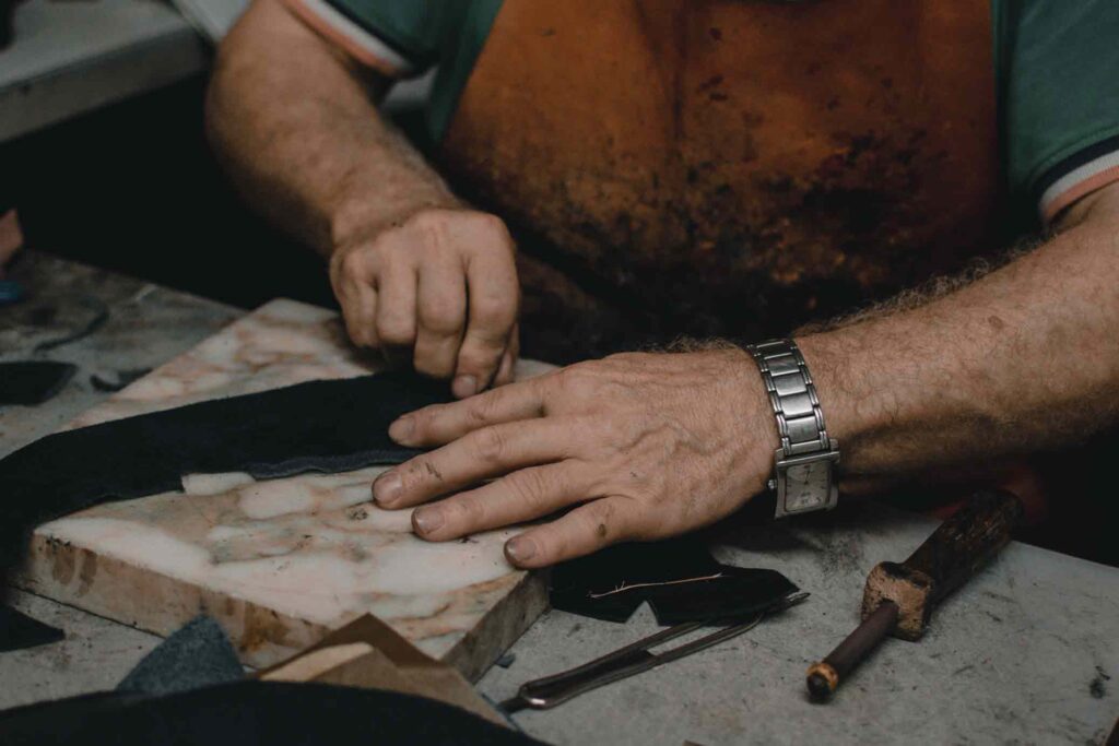 A man wearing a watch does leather working on a work bench. He wears a brown leather apron 