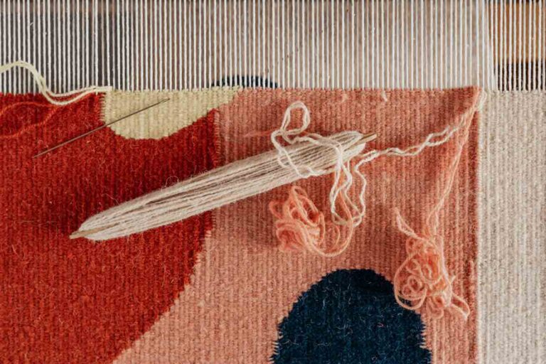 A close up image of a rug in the process of being woven, with pink, red and blue yarn. The rug is handmade to support local designers