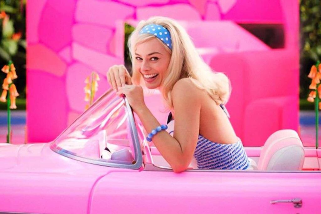 Margot Robbie as Barbie riding in a pink convertible smiling at the camera.