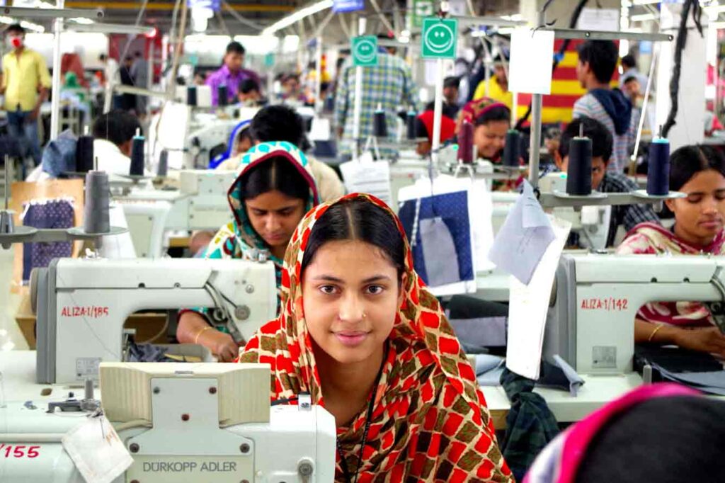 A woman sews at a machine wearing an orange head scarf and smiling.